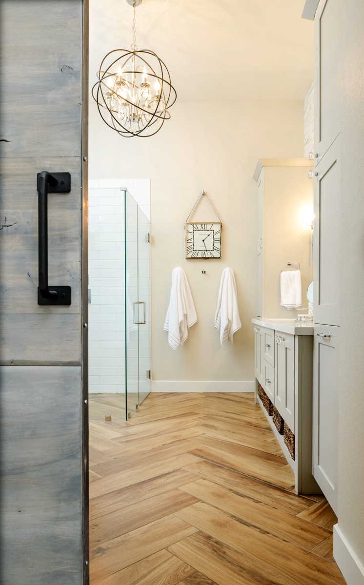 Looking through a barn door into a bathroom with distressed flooring and light grey cabinetry.