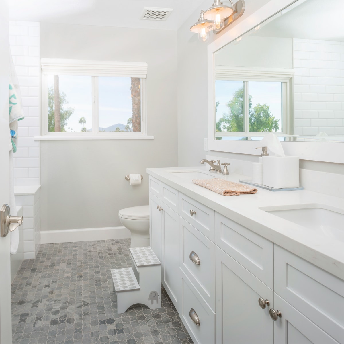 A white shaker-style bathroom vanity with grey tile flooring and a children's stepstool in front of a sink.
