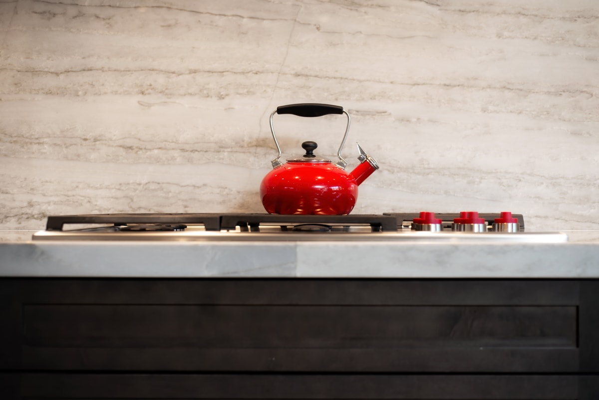 Bright red kettle sitting on a stove in front of a neutral stone backsplash.