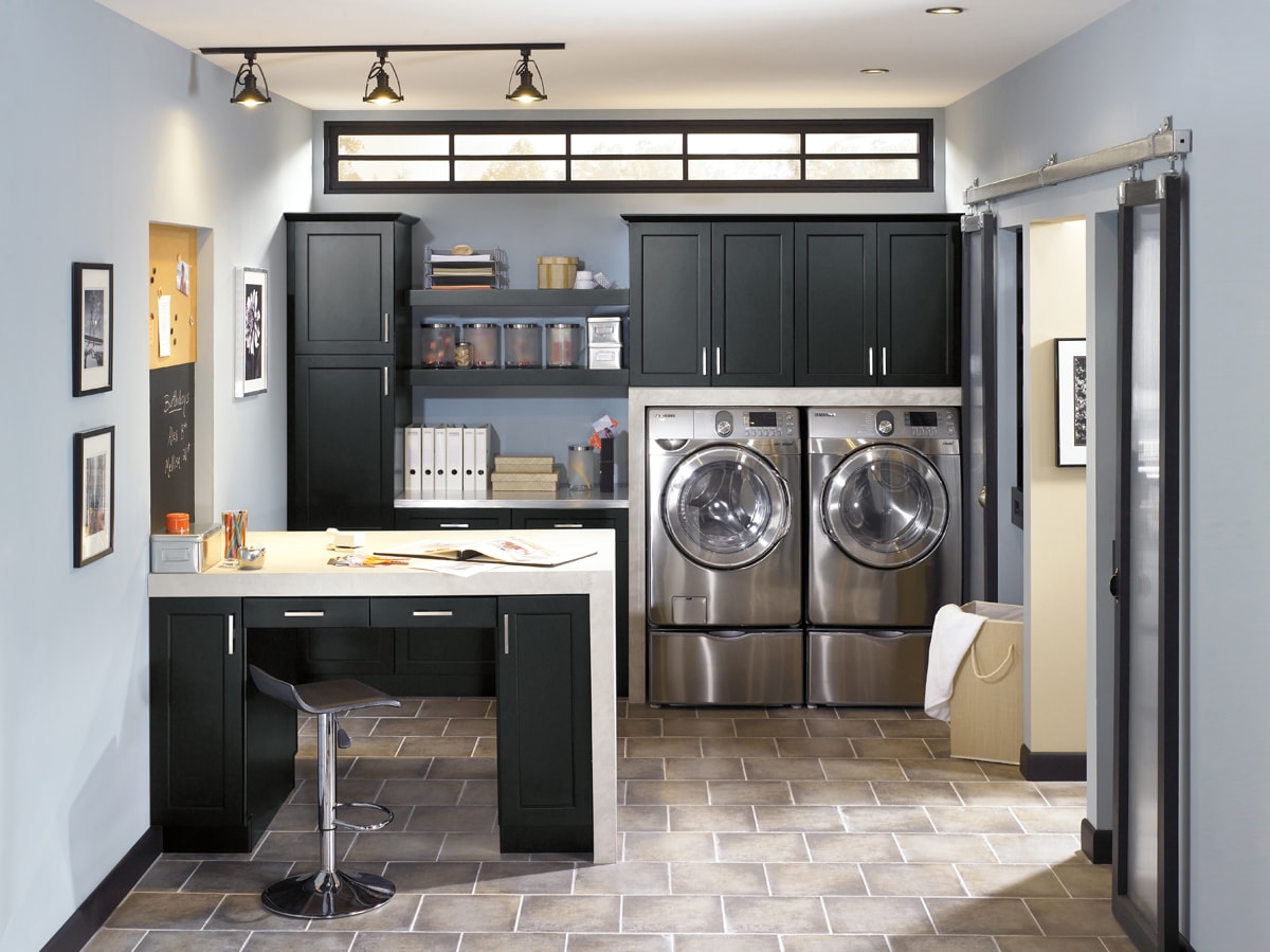 Laundry room with black cabinets abouve the washer and dryer. Left side of the room has a tall desk with a stool.